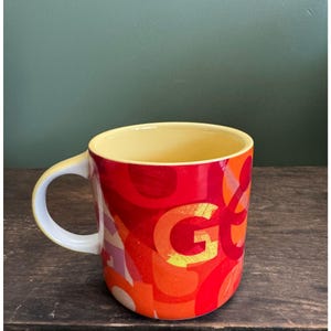 May include: A ceramic mug with a yellow interior and a white handle. The exterior features a vibrant design with the letter "G" in large orange letters, set against a background of red, orange, and yellow brushstrokes.