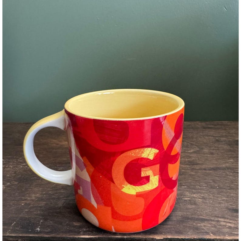 May include: A ceramic mug with a yellow interior and a white handle. The exterior features a vibrant design with the letter "G" in large orange letters, set against a background of red, orange, and yellow brushstrokes.
