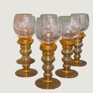 May include: A set of four clear glass goblets with etched floral patterns on the bowls. Each goblet features a distinct amber-colored stem with a stacked, rounded, ball-like design and a wide, flat circular base for stability.