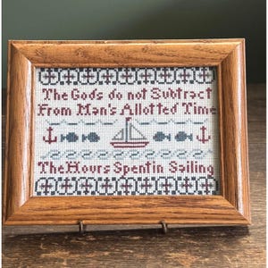 May include: A framed cross-stitch artwork with a nautical theme. The text reads "The Gods do not Subtract From Man's Allotted Time The Hours Spent in Sailing." The design includes a sailboat, anchors, and fish, all in shades of red, blue, and gray.