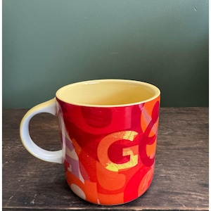May include: A ceramic coffee mug with a yellow interior and a white handle. The exterior features a vibrant design of red, orange, and yellow abstract shapes and the letter "G". The mug sits on a wooden surface.