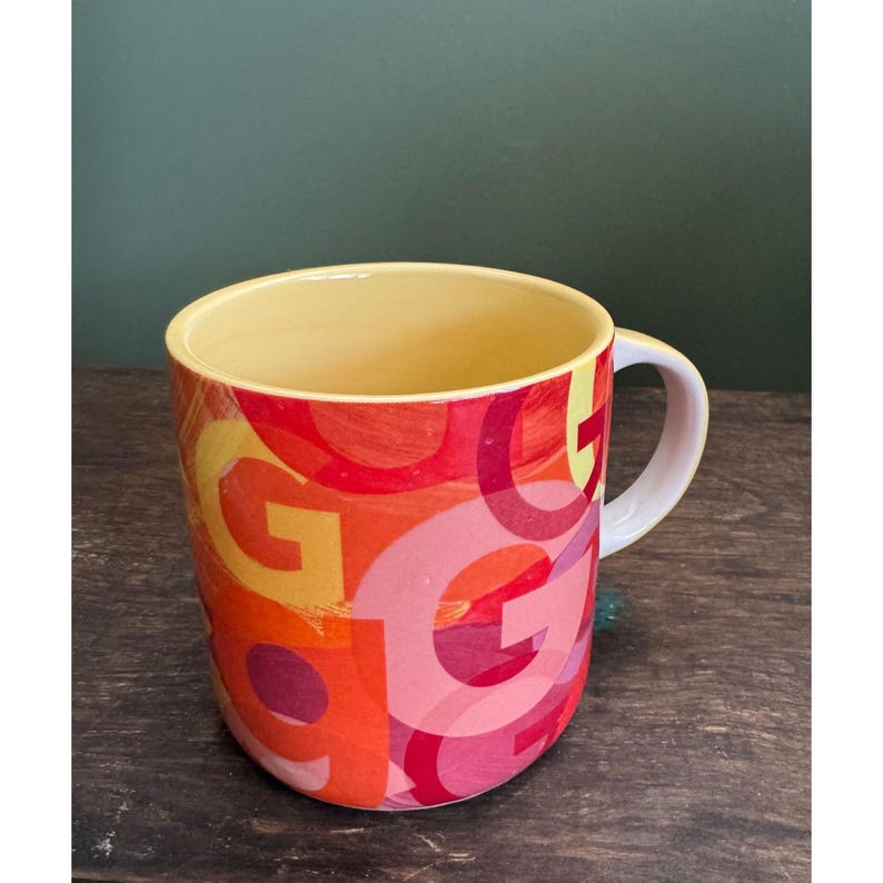 May include: A ceramic mug with a white handle and a yellow interior. The exterior features a colorful design of overlapping "G" letters in shades of red, orange, and pink. The mug sits on a dark wooden surface.