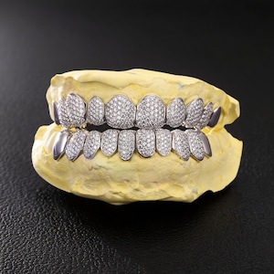 May include: A set of silver-colored dental grills, encrusted with numerous small, sparkling stones, displayed on a yellow dental mold. The grills are designed to cover the upper and lower teeth, showcasing a luxurious and eye-catching aesthetic.