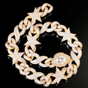 Può includere: Collana a catena color oro con ciondoli a forma di stella e occhio, incastonata con cristalli scintillanti. La collana presenta un design a maglie audace e grosso, creando un accessorio lussuoso e accattivante. I ciondoli a stella aggiungono un tocco celeste.
