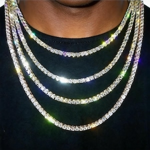 Op de afbeelding: Close-up van meerdere goudkleurige tenniskettingen met sprankelende, heldere edelstenen. De kettingen zijn gelaagd, wat een gedurfde uitstraling creëert. De achtergrond is effen zwart, waardoor de schittering van de sieraden wordt benadrukt.