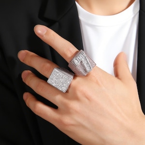 May include: Two square-shaped rings with a silver finish and encrusted with small, sparkling crystals. One ring is on the index finger, and the other is on the middle finger. The rings are a statement piece of jewelry.