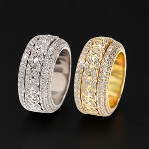 May include: Two rings, one silver and one gold, are displayed against a black background. Both rings feature a chain-link design in the center, surrounded by a row of small, sparkling stones. The gold ring has a yellow hue.