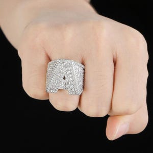 Könnte beinhalten: Ein silberfarbener Ring mit dem Buchstaben "A", besetzt mit kleinen, funkelnden Steinen. Der helle Ring steht im Kontrast zu dem dunklen Hintergrund. Ein auffälliges Schmuckstück.