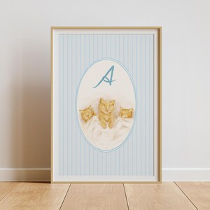 Puede incluir: Impresión artística enmarcada con tres gatitos naranjas durmiendo en una manta blanca dentro de un marco ovalado. El fondo tiene rayas verticales azul claro y una letra "A" azul en la parte superior.