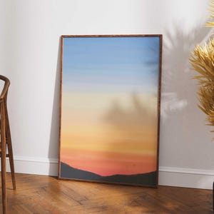 Puede incluir: Una impresión artística enmarcada que representa un paisaje de puesta de sol con un degradado de colores que va del azul al naranja y al rojo. La obra de arte se exhibe sobre un suelo de madera, apoyada en una pared blanca. A la izquierda se ve una silla de madera.
