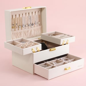May include: A cream-coloured jewellery box with a hinged lid, multiple tiered trays, and a bottom drawer. The interior features hooks for necklaces, a gathered pocket, and various compartments lined with beige fabric. Gold-tone metal hardware is visible.