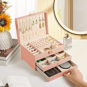 May include: A pink jewelry box with multiple compartments and drawers. The box is open, revealing various necklaces, earrings, rings, and bracelets. The box has a gold-colored handle and clasp. The interior is lined with a light-colored fabric.