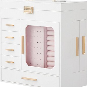 May include: A white jewelry box with multiple compartments, drawers, and a handle. The box features gold-colored hardware, including drawer pulls and a clasp. The interior is lined with pink fabric, and the box has a clear glass door.