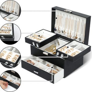 May include: A black jewellery box with a white interior, featuring a hinged lid with necklace hooks, a removable tiered tray for rings and earrings, and a bottom drawer with multiple compartments for organising various accessories and jewellery pieces.
