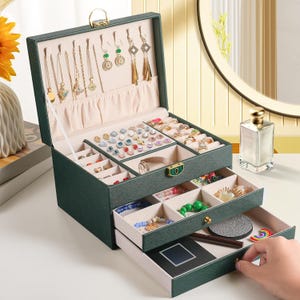 May include: A dark green jewelry box with multiple compartments and drawers. The top section displays necklaces and earrings, while the drawers hold rings, bracelets, and other accessories. The box is open, revealing various jewelry pieces.