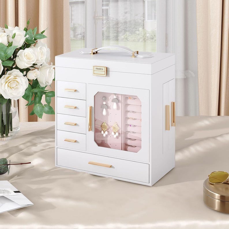 May include: A white jewelry box with multiple drawers and a glass-fronted compartment. The box has gold-colored handles and accents. The top features a white handle and a gold-colored clasp. The jewelry box is sitting on a light-colored surface.