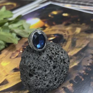 May include: A silver-toned ring with an oval, deep blue gemstone. The ring is set on a dark, textured rock, with a blurred background. The gemstone is faceted, catching the light. The ring's band has a detailed, textured design.