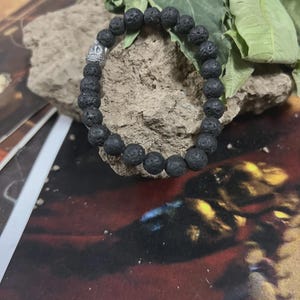May include: Black lava rock bead bracelet with a silver-colored accent bead. The bracelet is made of round, porous beads. The bracelet is displayed on a rock with green leaves in the background. The bracelet is a piece of jewelry.