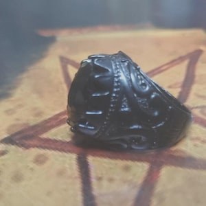 May include: A black ring with intricate, raised detailing. The ring's surface is highly textured, with patterns and designs that create a complex visual appearance. The ring is set against a blurred background.