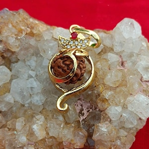 May include: A gold-colored pendant with a brown, textured center, adorned with a red gemstone and clear crystals. The pendant is set against a light-colored, textured background with a red backdrop.