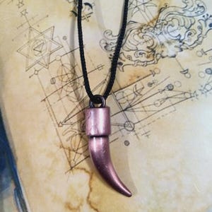 Puede incluir: Collar con colgante en forma de cuerno de color cobre, colgado de un cordón negro. El fondo es un mapa vintage con diseños geométricos.
