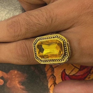 May include: A gold-coloured ring featuring a large, rectangular, yellow gemstone. The ring has a decorative border and a textured band. The ring is on a finger.