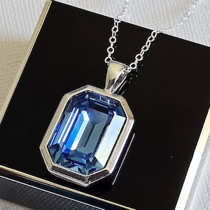 May include: A silver pendant necklace with a large, rectangular, faceted blue gemstone. The gemstone is set in a silver bezel and hangs from a delicate silver chain. The necklace is displayed on a black surface.