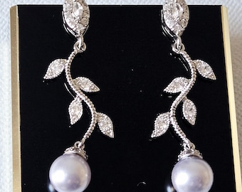 Könnte beinhalten: Elegante silberne Ohrringe mit Blattdesign und einer hängenden Perle. Die Ohrringe haben eine markiseförmige Oberseite und sind mit funkelnden Kristallen verziert. Die Perlen sind in einem sanften, hellen Lila gehalten.