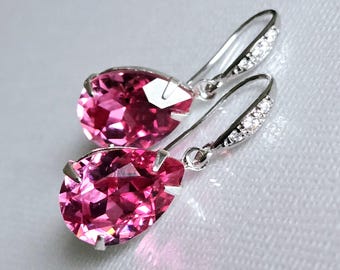 May include: A pair of drop earrings featuring large, faceted, teardrop-shaped pink gemstones. The silver-toned ear wires are adorned with small, clear crystals. The earrings are set against a soft, white background.