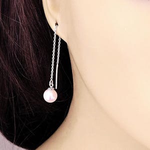 May include: A single silver chain earring with a dangling, light pink pearl. The earring is shown in wear, with the chain extending from the ear. The pearl is round and has a subtle sheen.