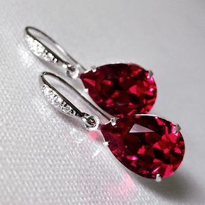 May include: A pair of silver-toned drop earrings with large, faceted, teardrop-shaped red gemstones. The earring hooks are adorned with small, clear crystals, adding sparkle. The earrings are displayed on a white, textured surface.