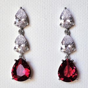 Peut inclure: Boucles d'oreilles pendantes à trois niveaux. Les deux premiers niveaux présentent des pierres claires en forme de poire, tandis que le niveau inférieur présente une pierre rouge foncé en forme de poire. Les boucles d'oreilles sont serties dans un métal argenté.