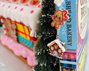 May include: A festive scene featuring a gingerbread house, a miniature Christmas tree, and a charm necklace with gingerbread man and house pendants. The gingerbread house has a pink and white roof with colorful details. The necklace hangs near a book titled "Gingerbread Bakery".