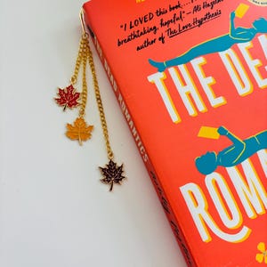 May include: A gold-colored metal bookmark with three maple leaf charms in red, orange, and brown, hanging from a chain. The bookmark is placed in a book titled "The Dead Romantics" with a bright orange cover.