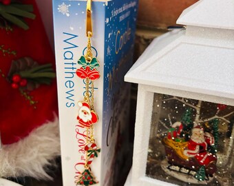 May include: A gold-colored Christmas-themed bookmark with red, green, and white enamel charms. The charms include bells, Santa Claus, a candy cane, and a Christmas tree. The bookmark is inserted into a book with the title "Love at Christmas".
