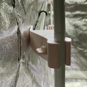 Könnte beinhalten: Nahaufnahme eines beigefarbenen Kunststoffclips, der an einer silbernen, reflektierenden Oberfläche und einer weißen Stange befestigt ist. Der Clip hat einen Hebel und dient dazu, die Stange zu halten. Der Hintergrund ist ein reflektierendes Material.