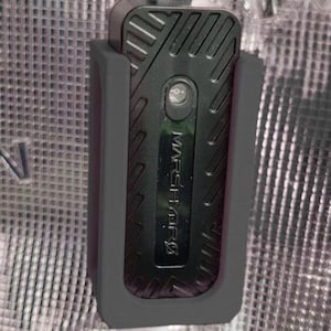 Puede incluir: Un dispositivo electrónico negro con la palabra "MARSHMFS" impresa, dentro de un soporte gris. El dispositivo tiene una superficie texturizada con un pequeño botón circular y un diseño acanalado.