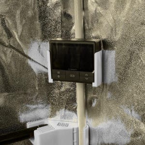 Könnte beinhalten: Ein schwarzer AC Infinity-Controller mit digitaler Anzeige ist an einer weißen Halterung befestigt. Der Controller hat mehrere Tasten und einen kleinen Bildschirm. Die Halterung ist an einer weißen Stange vor einem silbernen Hintergrund befestigt.