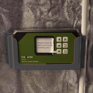 Könnte beinhalten: Ein TrolMaster TCS-1 Tent-X Steuerungssystem in einer grauen Kunststoffhalterung. Das grüne und schwarze Gerät verfügt über einen digitalen Bildschirm mit dem Text SENSOR MUST BE INSTALLED TO USE CONTROLLER! und sechs weiße Tasten.