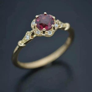 Op de afbeelding: Een gouden ring met een ronde, dieprode edelsteen in het midden, omringd door kleinere, heldere stenen. De band en zetting van de ring hebben een sierlijk, vintage-geïnspireerd ontwerp. De ring staat tegen een donkere achtergrond.