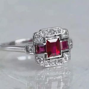 May include: A silver ring with a central square-cut ruby, two smaller rubies, and a halo of small diamonds. The ring has an art deco design. The ring's design is reminiscent of the 1920s.