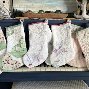 May include: A collection of six quilted Christmas stockings hanging in a row. Each stocking features unique patchwork designs in various colors including green, white, pink, and brown, with intricate stitching patterns and fabric textures throughout.