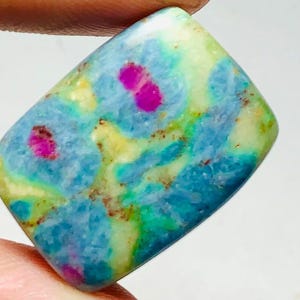 May include: A rectangular gemstone with a polished surface. The stone features a mix of colours, including blue, green, yellow, and pink. The pink areas appear as small, irregular spots within the stone's overall pattern. The gemstone is held between fingers.