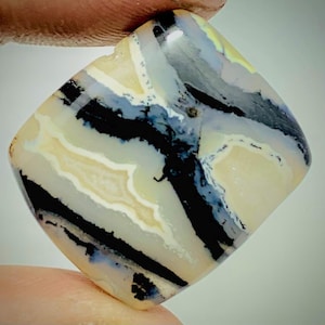 Op de afbeelding: Een vierkant geslepen edelsteen met een uniek patroon van zwarte, witte en lichtgele banden. De steen wordt tussen de vingers gehouden, waardoor het gepolijste oppervlak en de natuurlijke kleurvariaties worden benadrukt. Het ontwerp van de edelsteen doet denken aan abstracte kunst.