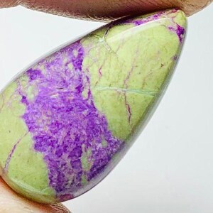 Peut inclure: Une pierre précieuse polie en forme de goutte d'eau. La pierre présente une base vert pâle, recouverte de taches violettes profondes. De fines veines de couleur claire traversent la surface, ajoutant de la texture.