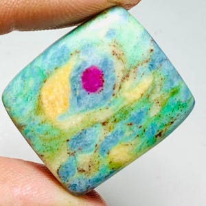 Könnte beinhalten: Ein quadratischer Edelstein mit einer lebendigen Farbpalette. Der Stein weist einen zentralen fuchsiafarbenen Punkt auf, der von konzentrischen Mustern in Hellblau, Gelb und Grün umgeben ist. Der Stein wird von einem Finger gehalten.