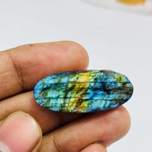 Könnte beinhalten: Ein ovaler Labradorit-Edelstein mit polierter Oberfläche. Der Stein zeigt ein lebendiges Farbspiel mit schillernden Blau-, Grün- und Gelbtönen. Der Edelstein wird in einer Hand gehalten, was seine natürliche Schönheit und einzigartigen Muster zeigt.
