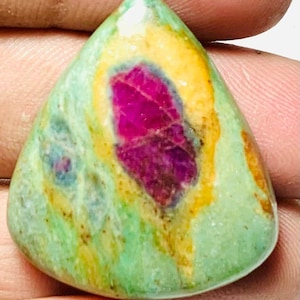 May include: A polished, teardrop-shaped gemstone with a vibrant colour palette. The stone features a central, deep magenta core surrounded by a yellow and orange halo, set against a backdrop of green and blue hues. The gemstone is smooth and appears to be natural.