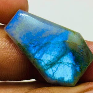 Könnte beinhalten: Ein polierter Labradorit-Edelstein mit rechteckiger Form und abgeschrägten Kanten. Der Stein zeigt ein lebendiges Farbspiel mit Blau- und Grüntönen, die über seine Oberfläche wirbeln. Der Edelstein wird in einer Hand gehalten.