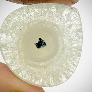 May include: A polished, off-white gemstone with a distinctive pattern. The stone features a central dark blue-black inclusion, encircled by rings of varying opacity. The outer edge has a textured, crystalline appearance.